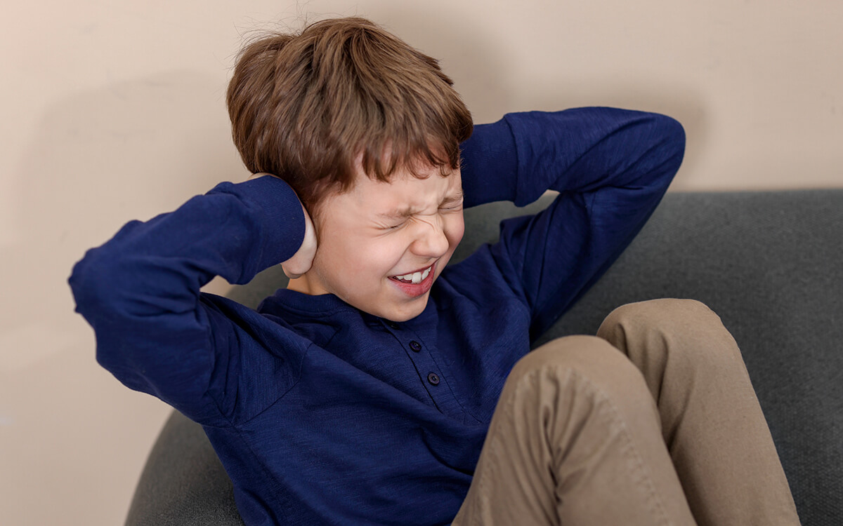 Child covering his ears while sitting on a couch, showing signs of distress and overwhelm.