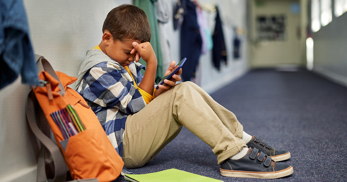 Young student sitting on the floor in a school hallway, looking distressed while holding a smartphone beside his backpack.